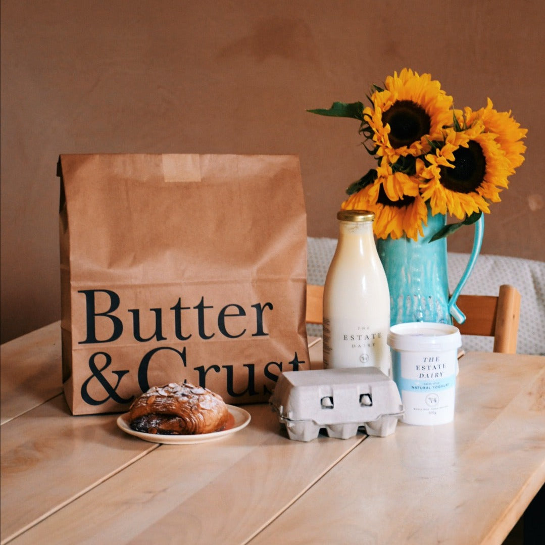 A paper bag with 'Butter & Crust' branding, a bottle of milk, a carton of eggs, a container of yoghurt, a croissant, and a vase with sunflowers on a wooden table.