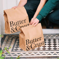 Two people holding brown paper bags with 'Butter & Crust' printed in black. The bags are placed on a doorstep with a black and white tiled floor visible.