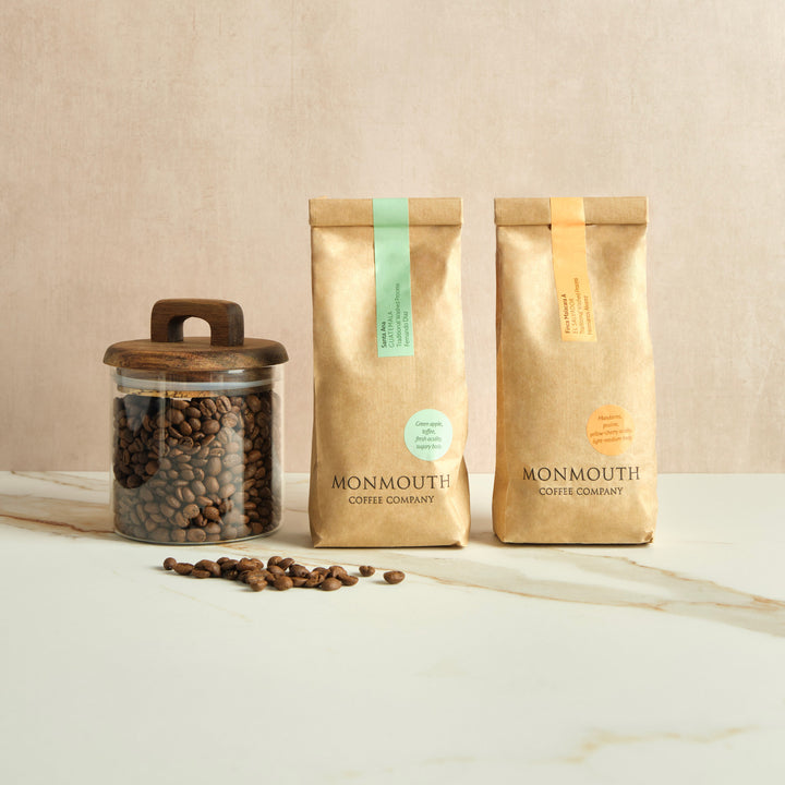 Two Monmouth Coffee Company bags and a jar of coffee beans on a marble surface with a beige background.