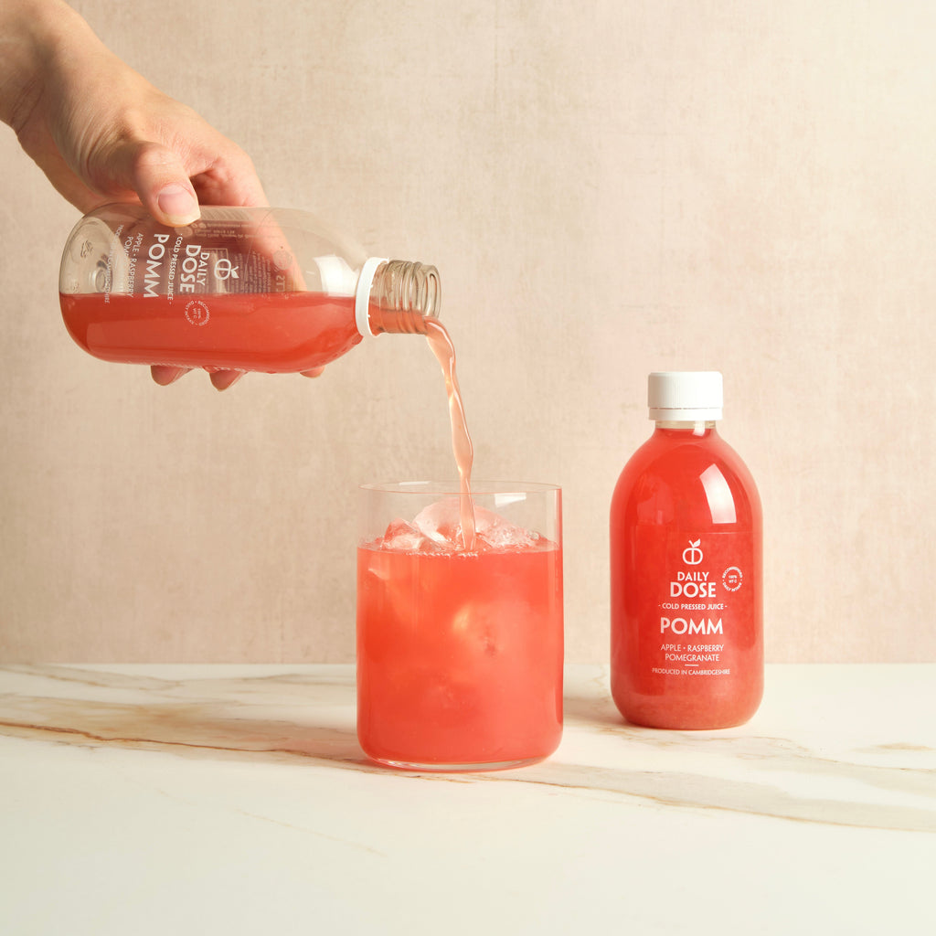 Hand pouring pink juice from a bottle labeled "Daily Dose Pomm" into a glass on a marble surface with a beige background.