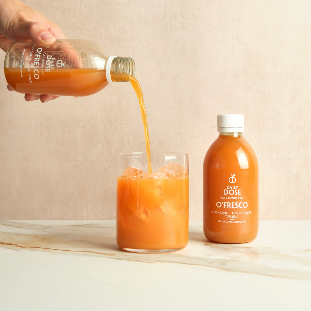 Person pouring carrot juice from a bottle labeled 'Daily Dose O'Fresco' into a glass on a marble surface with a beige background.