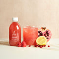 Bottle labeled "Daily Dose Pomm" and a glass of pink juice with pomegranate, raspberries, and lemon on a marble surface with a beige background.
