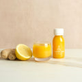 Bottle labeled 'Daily Dose Turmeric Shot' and glass of it with a ginger root and lemon on a marble surface with a beige background.