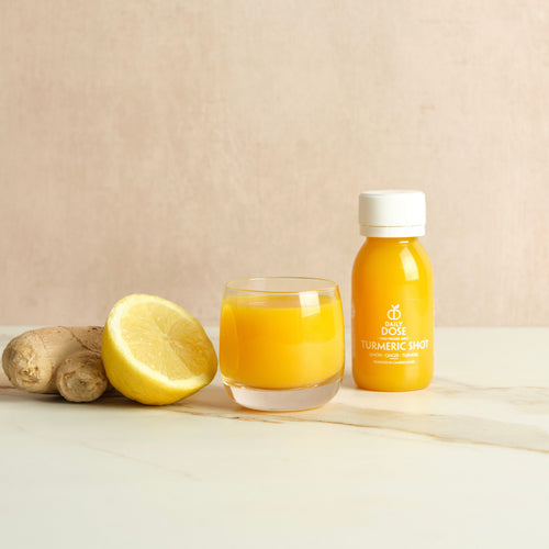 Bottle labeled 'Daily Dose Turmeric Shot' and glass of it with a ginger root and lemon on a marble surface with a beige background.