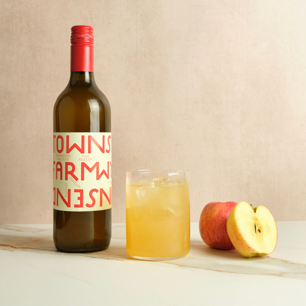 Bottle of farm pressed apple juice accompanied by a filled glass of apple juice and an apple on a plain background.