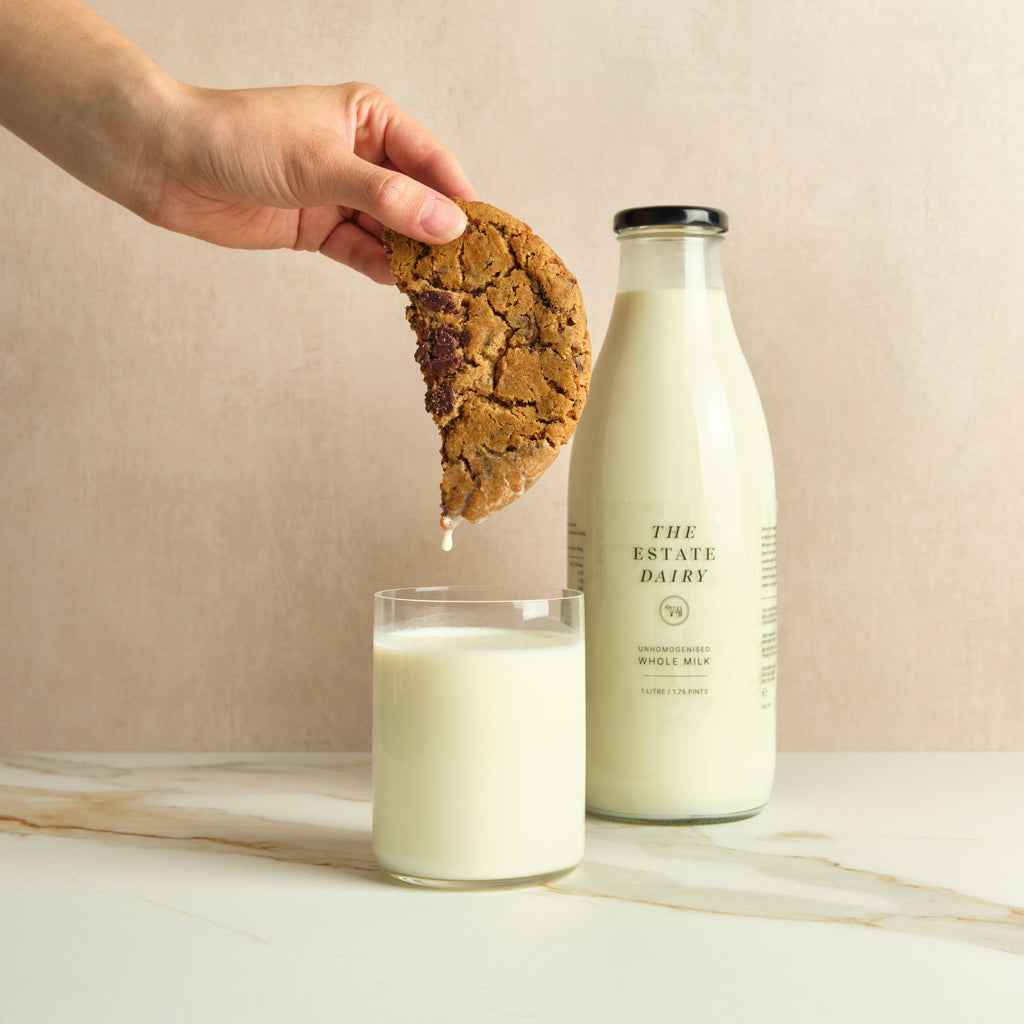 Hand dipping a cookie into a glass of milk next to a bottle of milk with 'The Estate Dairy' branding on a plain background.