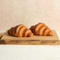 Two plain croissants on a wooden cutting board with a beige background.