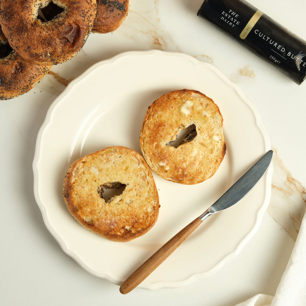 Buttered poppyseed bagels on a plate with a knife and a The Estate Dairy butter on a marble surface.