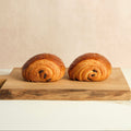 Two chocolate croissants on a wooden cutting board with a beige background.