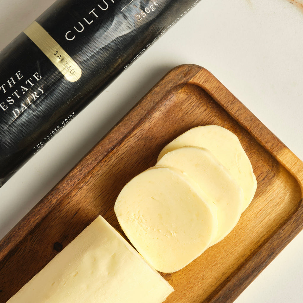 Sliced butter on a wooden cutting board next to The Estate Dairy salted cultured butter package. 