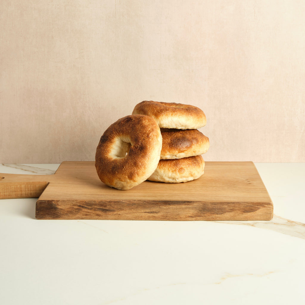 Four plain bagels stacked on a wooden cutting board against a beige background.