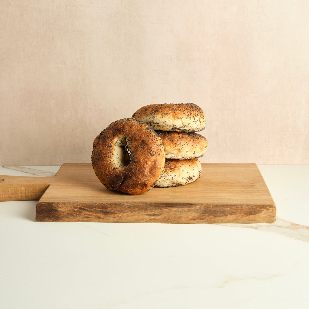 Four poppyseed bagels stacked on a wooden cutting board against a beige background.