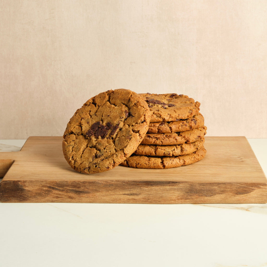 Six chocolate chip cookies on a wooden cutting board with a plain background.