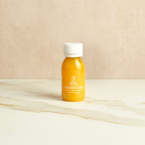 Small bottle with a white cap labeled 'Daily Dose Turmeric Shot' on a marble surface with a beige background.