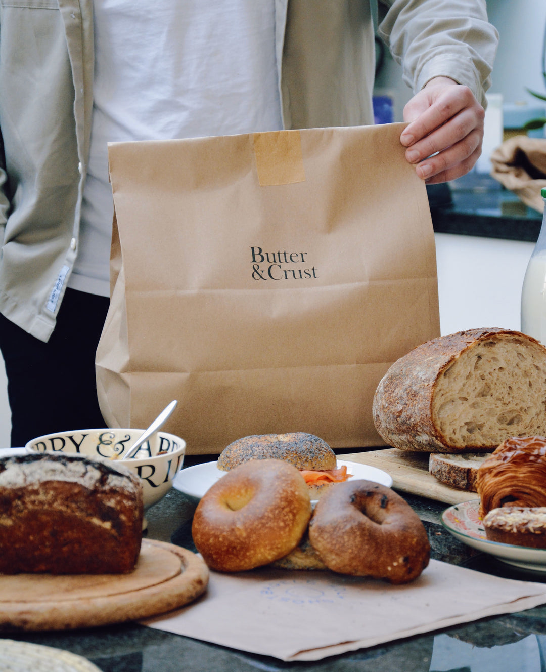 Hand holding Butter & Crust bundle with a selection of artisan breakfast goods int he foreground including: bagels, rye bread, sourdough and pastries.