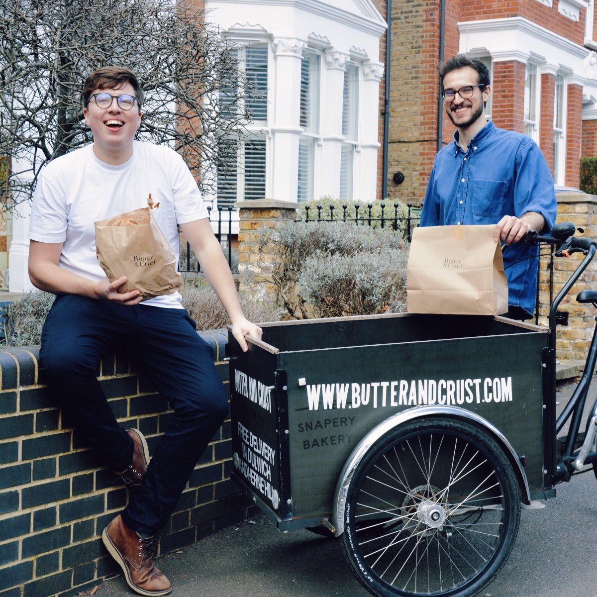 Butter & Crust co-founders Ollie (sitting on brick wall) and Nic (standing behind one of the branded Butter & Crust delivery bikes) each holding a Butter & Crust breakfast delivery bundle. 