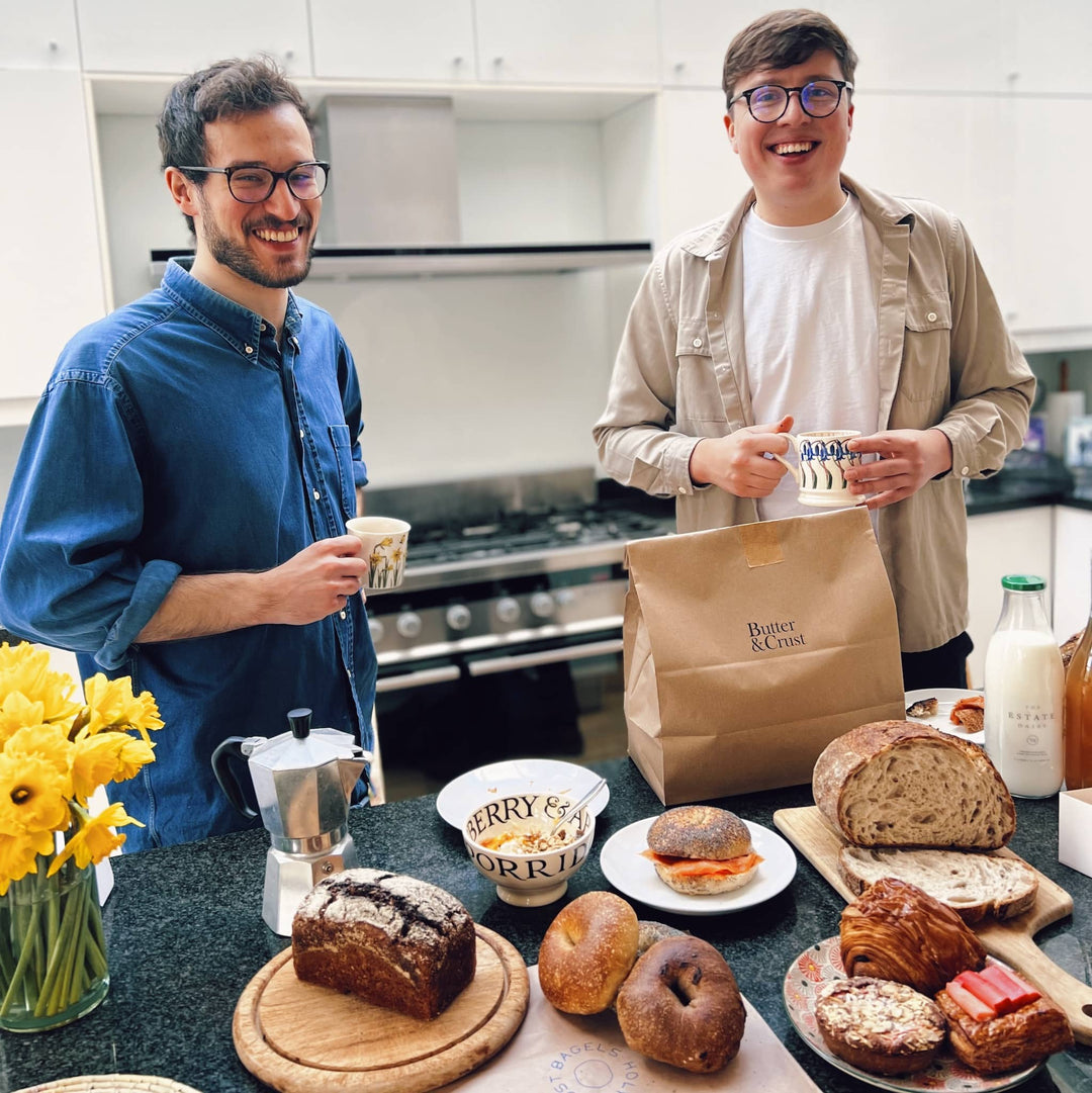Butter and Crust cofounders Ollie and Nic in a kitchen, holding a cup of coffee each, with a selection of the Butter and Crust breakfast goods including: buttermilk rye bread, bagels, pastries, the signature sourdough, granola, coffee, milk and a bunch of daffodils in the corner.