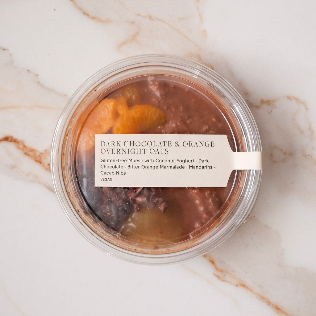 Round, labeled container of dark chocolate, mandarin, and coconut yoghurt overnight oats on a marble surface.