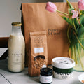 A breakfast bundle including a bottle of milk, a bag of granola, a carton of yogurt, a container of plum jam, and a bag labeled 'Butter & Crust', displayed on a table with pink tulips in a vase.