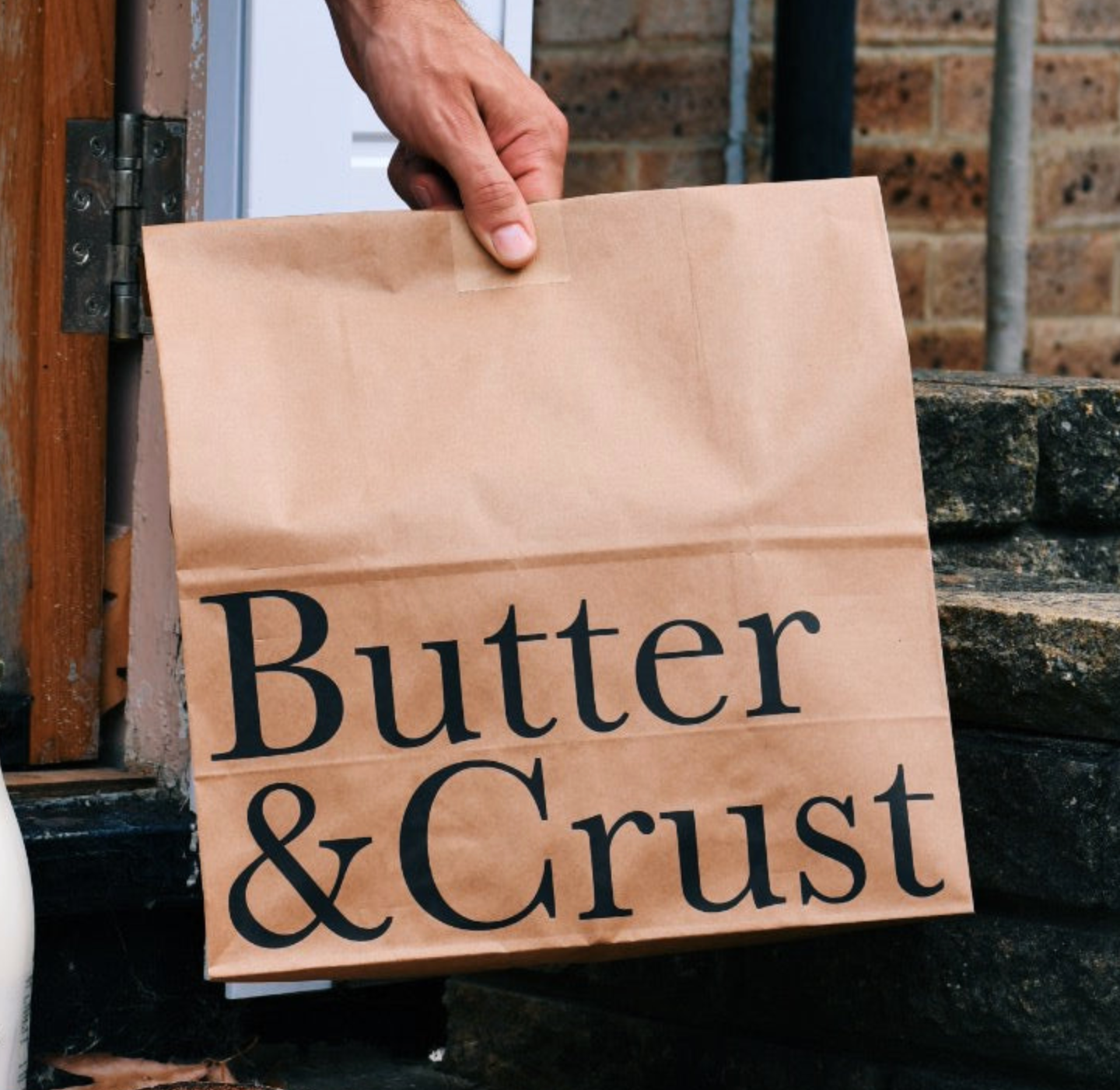 Person holding a brown paper bag with 'Butter & Crust' printed on it, outdoors.